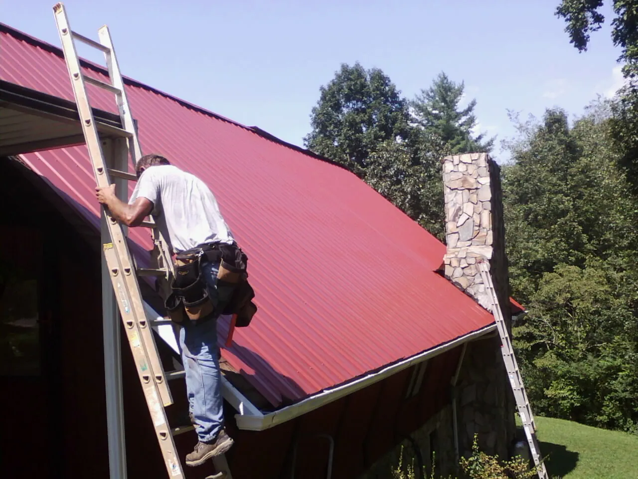 Roofer on a ladder beside a red metal roof and stone chimney — Soffit & Fascia Repair by Roofing Brothers in Gray Summit